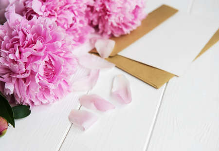 invitation card, craft envelope and pink peony flowers on a white wooden tableの写真素材
