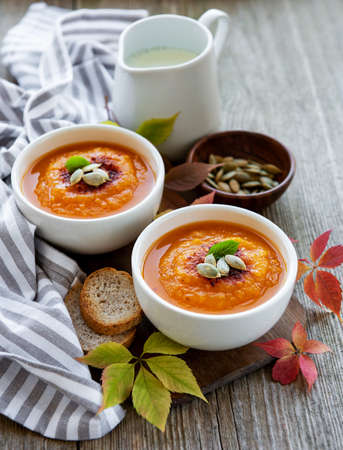 Bowls of pumpkin soup, fresh pumpkins and autumn leaves on rustic wooden background
