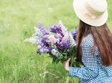 Back view g, viewirl in a summer field with a straw hat and a bouquet of lupins