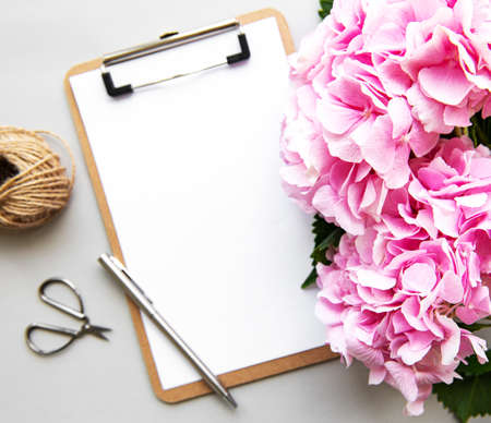 Home office desk workspace with blank paper clipboard, pink hydrangea flowers bouquet on whiteの写真素材