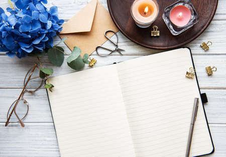 Home office desk workspace with blank notebook, blue hydrangea flower bouquet,  candles on white...