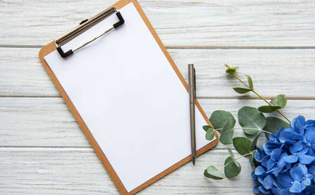 Home office desk workspace with blank paper clipboard, blue hydrangea flowers bouquet on white...
