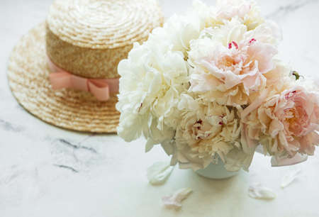 Straw hat on a table with peony flowers in vase at the windowの写真素材