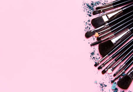 Different makeup brushes and broken eye shadow on pastel pink background. Flat lay, top view.