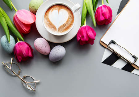 A cup of  coffee  over a gray background, white tulips and  Easter eggs and empty clipbiard.  Copy...
