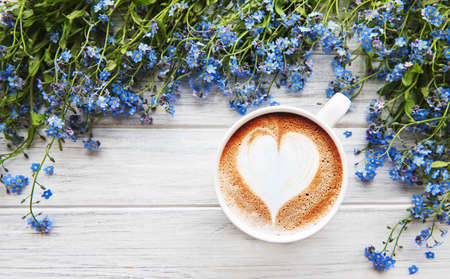 Blue forget-me-not flowers and a cup of hot coffee on a white wooden tableの写真素材