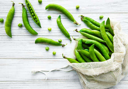 Reusable eco-friendly bag full of fresh pea pods on white wooden background