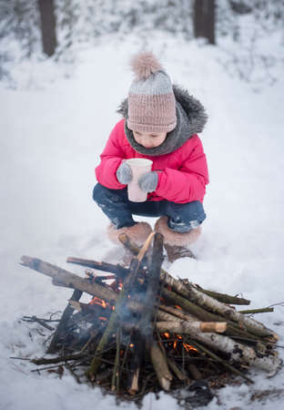 Child in winter. Little girl in winter in nature drink tea by the fireの写真素材