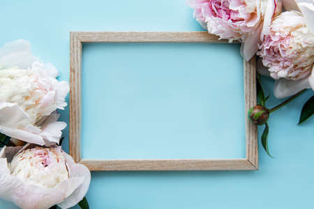 Wooden frame surrounded by beautiful pink peonies on a blue background, top view, copy space, flat lay. Mockup greeting card, invitations to a holiday or wedding. Bright summer flower concept.の写真素材