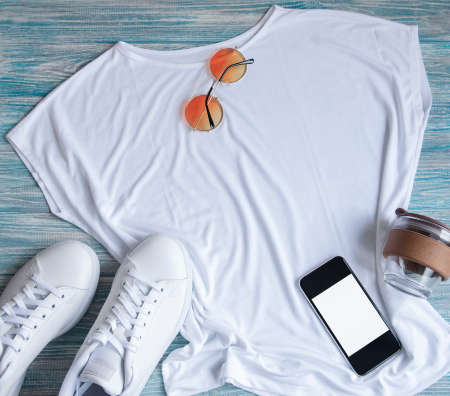 Stylish female clothes set. Woman outfit on blue wooden background. Blue jeans, white t-shirt, white sneakers and sunglasses. Flat lay, top viewの写真素材