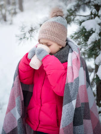 Little girl wrapped in a blanket drinking tea in the winter forestの写真素材