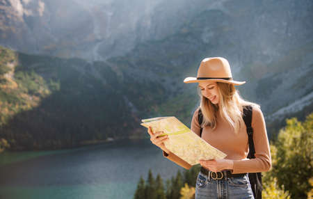 Wanderlust and travel concept. Stylish traveler girl in hat looking at map, exploring woods. Young oman with backpack traveling at lake in forest.の写真素材