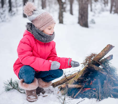 A little girl in a bright jacket plays in the winter snowy forest. The girl warms up by the fire.の写真素材