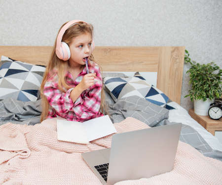Smiling happy little kid girl using laptop in wireless headphones in bed at home.の写真素材