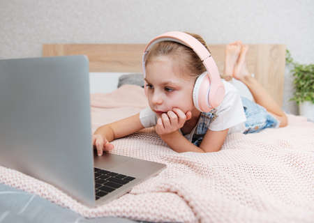 Smiling happy little kid girl using laptop in wireless headphones in bed at home.の写真素材