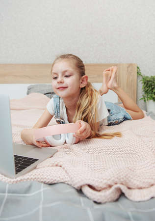 Smiling happy little kid girl using laptop in wireless headphones in bed at home.