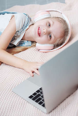 Smiling happy little kid girl using laptop in wireless headphones in bed at home.の写真素材