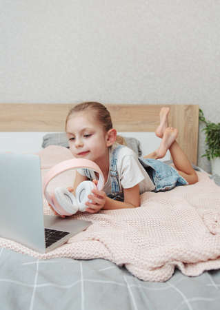 Smiling happy little kid girl using laptop in wireless headphones in bed at home.の写真素材
