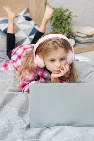 Smiling happy little kid girl using laptop in wireless headphones in bed at home.の写真素材