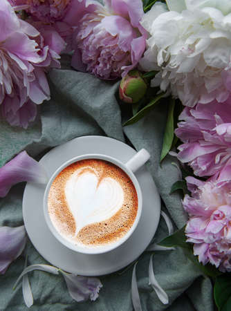 Flat lay cup of coffee with heart shape pattern surrounded with light pink peony flower petals on green textile backgroundの写真素材