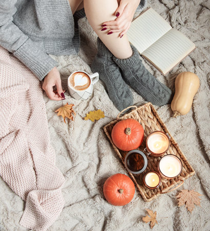 A girl in a sweater and knitted socks sits on a blanket. Cozy autumn concept. Pumpkins and candles on a tray near the girl.の写真素材