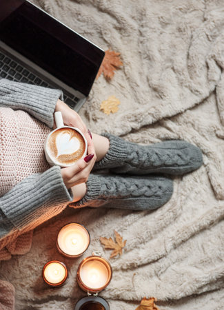 Cozy home, woman covered with warm blanket, drinks coffee and works on a laptop. Relax, carefree,...