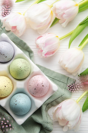 Holder  with Easter eggs painted pastel colors and pink tulips on a white wooden background. Top...