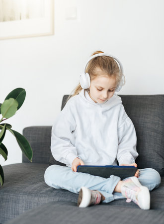 Little girl  lying on the couch with an electronic tablet listens to music in white headphones. Using wireless devices for education.の写真素材