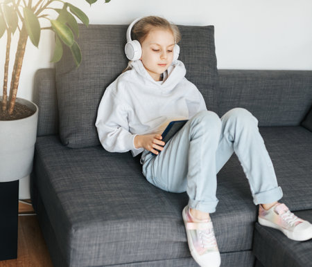 Little girl  lying on the couch with an electronic tablet listens to music in white headphones. Using wireless devices for education.の写真素材