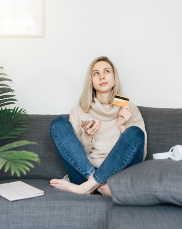 Young woman holding smartphone and banking credit card, involved in online mobile shopping at home, happy female shopper purchasing goods or services in internet store.の写真素材