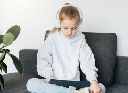 Little girl  lying on the couch with an electronic tablet listens to music in white headphones. Using wireless devices for education.の写真素材