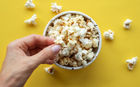 A woman's hand takes popcorn from a cup.  Yellow background.