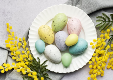 A selection of painted Easter eggs in pastel shades are elegantly displayed on a plate, accented with fresh yellow mimosa blooms.の写真素材