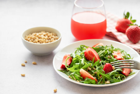 A vibrant plate of strawberry arugula salad, garnished with toasted pine nuts, presented on a white plate, set against a light grey backdrop. The freshness of the greens and the ripeness of the strawberries suggest a healthy, nourishing meal option.の写真素材