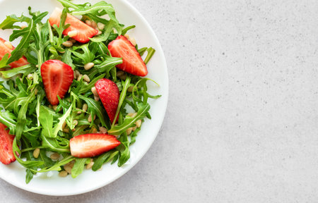 A vibrant plate of strawberry arugula salad, garnished with toasted pine nuts, presented on a white plate, set against a light grey backdrop. The freshness of the greens and the ripeness of the strawberries suggest a healthy, nourishing meal option.の写真素材