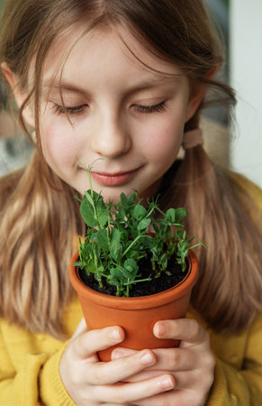 A child, dressed in a vibrant yellow sweater, delicately holds a terracotta pot with a green plant. She looks at the plant with a gentle smile and an apparent affection that suggests a connection with nature. The soft, natural light gives a warm atmosphere to the moment, capturing her subtle expression of joy and curiosity.の写真素材
