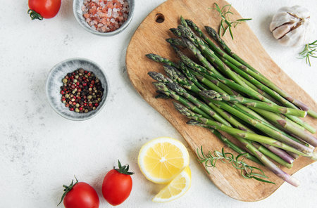 A bundle of fresh asparagus is laid out on a wooden cutting board alongside sliced lemons, robust tomatoes, garlic, and a mix of seasoning. The arrangement suggests the preparation of a healthy, flavorful meal, with the ingredients fresh and prominently displayed against a light background.の写真素材