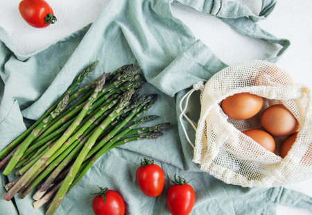A bunch of fresh asparagus lies next to a reusable mesh bag filled with brown eggs, accented by ripe cherry tomatoes on a light-colored surface with a blue cloth. The setting suggests preparation for a healthy, home-cooked meal.の写真素材