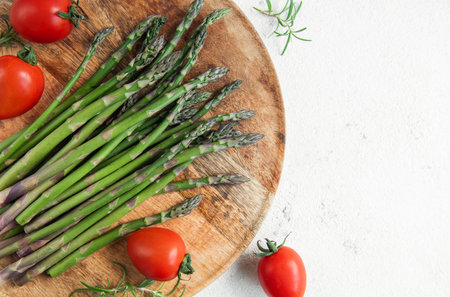 A collection of vibrant green asparagus spears are neatly arranged on a round wooden cutting board, accompanied by bright red cherry tomatoes. The arrangement is placed against a stark white backdrop, highlighting the freshness and colors of the produce. The scene suggests preparation for a healthy meal, with a focus on fresh vegetables.の写真素材