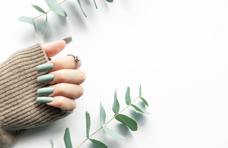 A delicate hand with styled green nails is elegantly positioned next to soft eucalyptus leaves. The hand is partially covered by a cozy sweater, creating a serene, minimalist aesthetic.の写真素材