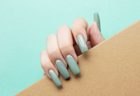A hand with elegantly manicured long green nails grips a brown envelope, contrasting beautifully with the light blue backdropの写真素材