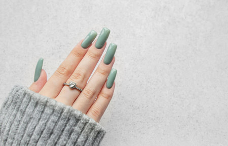 A cozy hand with long, stylish green nails features a delicate ring, elegantly resting on a soft gray sweater against a light background, creating a tranquil atmosphere.の写真素材