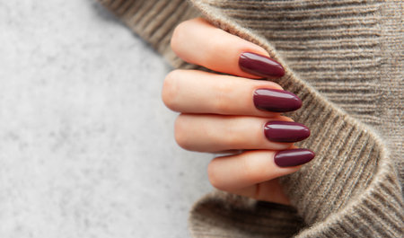 A well-manicured hand showcases dark burgundy nails while holding a cozy knitted sweater sleeve. The warm tones create an inviting, fashionable atmosphere.の写真素材