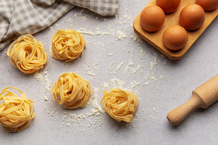Tagliatelle nests with flour, eggs and rolling pin placed on gray background, composing a beautiful food background for menu or recipe bookの写真素材