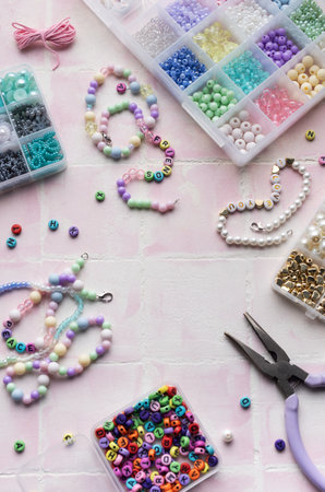 Crafting supplies including colorful beads, letter beads, string, clasps, and pliers are arranged on a pink tile surface, ready for jewelry makingの写真素材