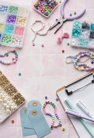Crafting materials including colorful beads, pliers, string, and letter beads are arranged on a pink tile background, alongside a clipboard and pens, creating a workspace for jewelry makingの写真素材