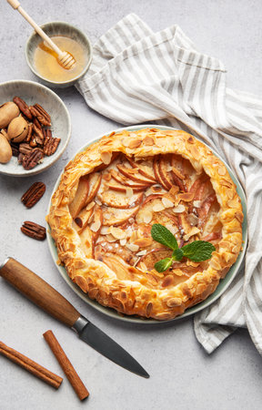 Freshly baked apple galette with almond flakes, cinnamon, and mint, surrounded by ingredients and kitchen utensilsの写真素材