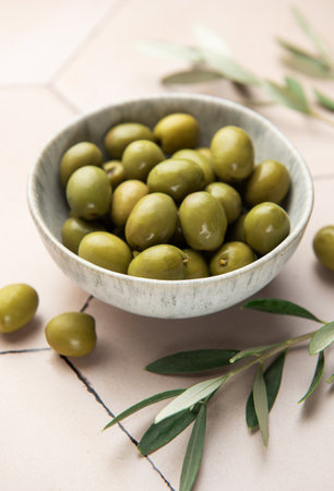 Green olives are served in a bowl alongside olive branches on a tiled surface, creating a mediterranean ambianceの写真素材