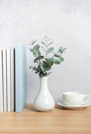 Modern home decor with eucalyptus branches in vase, books, and coffee cup on wooden desk against gray wall, creating cozy reading nookの写真素材