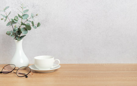 Eucalyptus branches in vase, coffee cup and eyeglasses sit on wooden desk against gray wall,...